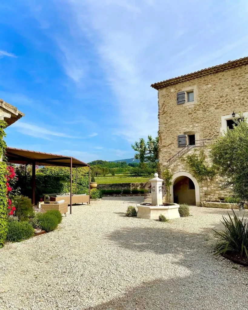 Cour provençale aménagée en gravier avec fontaine, végétation méditerranéenne et bâtisse ancienne en pierre