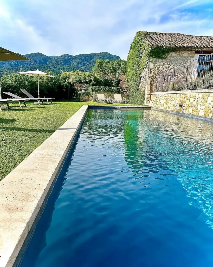 Piscine intégrée dans un jardin paysager avec pelouse, espace détente, bâtisse en pierre et vue sur le Luberon